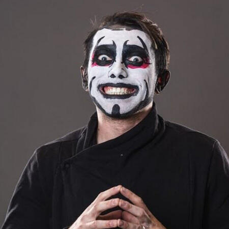 a photo of danhausen, a wrestler wearing black, white, & red full face paint and a black cloak-like shirt, with a wide grin and his fingers pressed together.