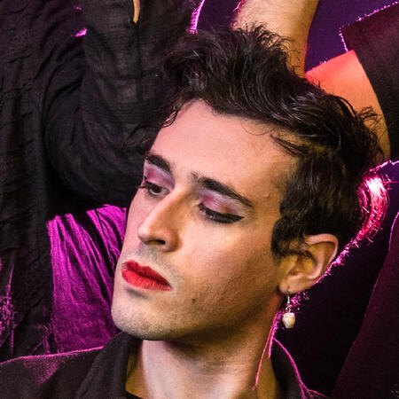 a photo of will wood, a white man with short curly black hair, wearing purple eyeshadow, red lipstick, and white earrings. he looks at something off-camera.